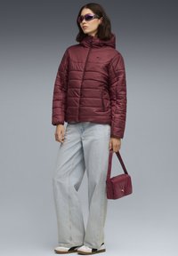 Bordeaux gewatteerde jas met een capuchon, voorzien van een rits aan de voorkant en zijzakken, gecombineerd met lichtblauwe wijde jeans en een bijpassende handtas.