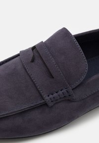 Mocassin en suède bleu marine avec un accent cousu au niveau du cou-de-pied, col surélevé et surface texturée. Intérieur lisse et design minimaliste.