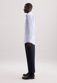 Camicetta azzurro chiaro a maniche lunghe con polsini a bottone, abbinata a pantaloni blu scuro e scarpe nere. Tessuto liscio, vestibilità slim e design minimalista.