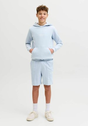 Garçon adolescent souriant, portant un sweat à capuche bleu clair et un short assorti, des chaussettes blanches et des baskets beige, debout les mains dans les poches.