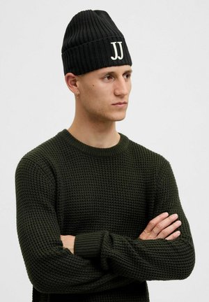 Black knitted beanie with a ribbed texture and white "JJ" logo on the front, worn with a green textured sweater.
