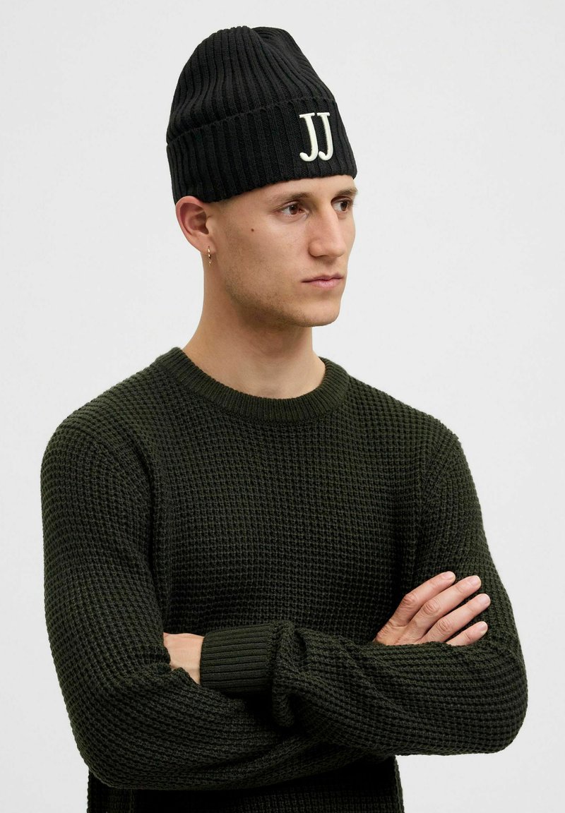 Gorro negro de punto con textura acanalada y logo blanco "JJ" en la parte frontal, combinado con un suéter texturizado verde.