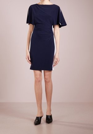 Frau trägt ein knie-umspielendes marineblaues Kleid mit flatternden Ärmeln, seitlichen weißen Einsätzen und schwarzen, spitz zulaufenden Mules, steht auf einem beigen Boden.