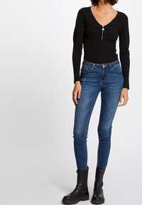 Svart långärmad topp med V-ringning och dragkedjedetalj fram, kombinerad med mörkblå slim fit-jeans och svarta ankelstövlar.