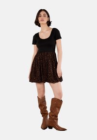 Giovane donna in piedi che indossa un top nero a maniche corte, una gonna con stampa leopardata e stivali marroni fino al ginocchio su uno sfondo semplice.