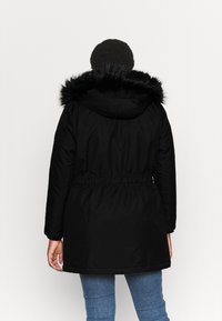 Parka preta com capuz de pelo sintético, cintura ajustada, mangas longas e textura suave. Parece quente e foi projetada para climas mais frios.
