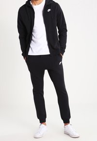 Felpa nera con zip, tasche frontali e logo Nike bianco, abbinata a pantaloni jogger coordinati e sneakers bianche. Tessuto morbido, design slim fit.