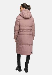 Kvinna iklädd en lång mauve-färgad quiltad huva, svarta leggings och svarta kraftiga stövlar, står vänd bort från kameran mot en vit bakgrund.