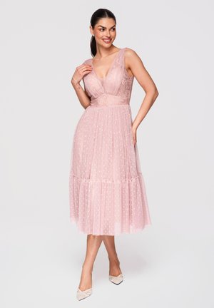 Kvinde iført ærmeløs pink midi-kjole i tyl og blonder med prikker, poserer med den ene hånd på hoften og den anden nær kravebenet.