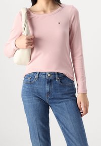 Długi rękaw, żeberkowy top w delikatnym różu, z okrągłym dekoltem i małym akcentem logo. Połączony z niebieskimi dżinsami i kremową torebką.