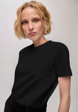 Femme blonde aux cheveux courts et ondulés portant un t-shirt noir à manches courtes sur un fond clair uni.