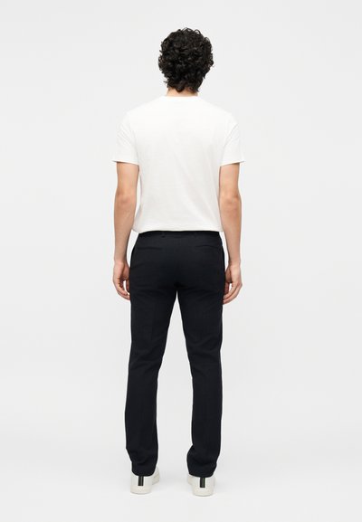 Άντρας που στέκεται με την πλάτη γυρισμένη, φορώντας ένα απλό λευκό κοντομάνικο μπλουζάκι, μαύρο παντελόνι και άσπρα αθλητικά παπούτσια με μαύρες ρίγες.