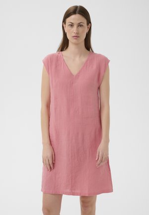 Žena stojaca pokojne v ružových šatách bez rukávov s výstrihom do V, dĺžky po kolená, na bielom jednofarebnom pozadí.