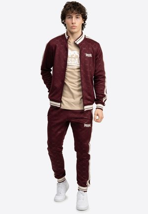 Bordeaux träningsdräkt med crèmefärgade ränder, bestående av en zip-jacka och matchande byxor. Toppdelen har en tryckt logotyp i vitt. Vita sneakers.