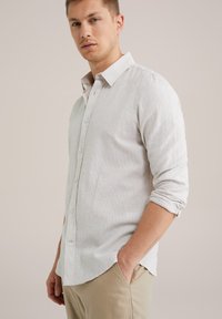 Camisa de botones gris claro con una tela texturizada, mangas largas remangadas en los puños y un dobladillo curvado. Llevada con pantalones beige.