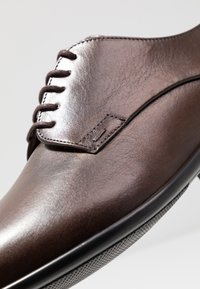 Scarpa da uomo in pelle marrone con una superficie liscia, progettata con chiusura con lacci e punta rotonda. Presenta un sottile dettaglio cucito.