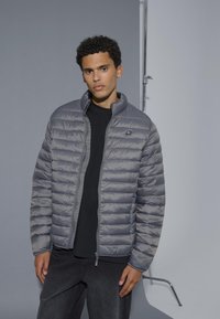 Grijze pufferjack met horizontale quiltpatroon, staande kraag en een ritsluiting. Gedragen over een zwart shirt, gecombineerd met donkere broek.