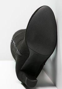 Svart mockakänga med en texturerad gummisula, som har en smal klack och sidodragkedje detaljer, vinklad mot en vit bakgrund.