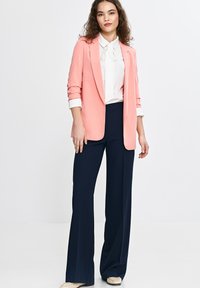 Jasnoróżowy marynarka z wyraźnymi ramionami, zestawiona z białą bluzką z krawatem i granatowymi spodniami o szerokich nogawkach.