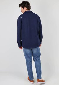 Chemise à manches longues bleu marine avec col, texture lisse, détails de boutons et un logo discret au dos, associée à un jean clair et des chaussures.