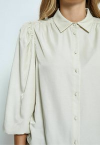 Light beige blouse with a collared neckline, button front, and gathered sleeves. The fabric has a smooth, slightly shiny texture.
