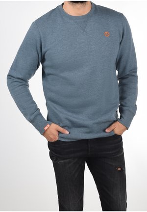 Homme portant un sweat-shirt bleu à manches longues avec un détail de logo orange sur la poitrine et le poignet, associé à un jean noir déchiré, mains dans les poches.