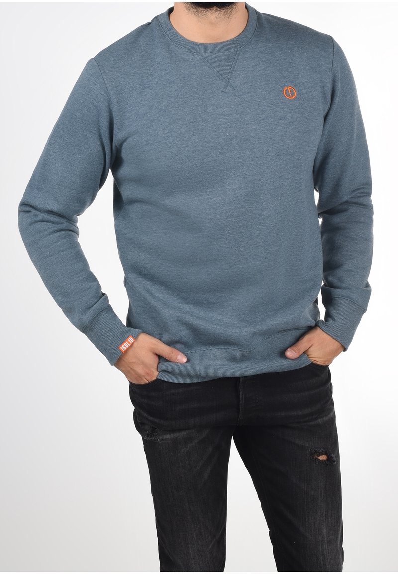 Homme portant un sweat-shirt bleu à manches longues avec un détail de logo orange sur la poitrine et le poignet, associé à un jean noir déchiré, mains dans les poches.