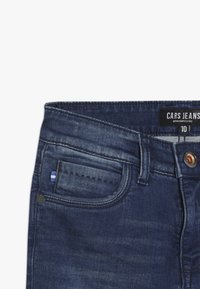 Tmavě modré džíny s úzkým střihem. Mezi prvky patří přední kapsa se modro-bílým pruhovaným detailem, měděný knoflík a značka.