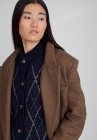 Blazer in misto lana marrone con una silhouette strutturata. Sovrapposto a un cardigan blu navy con un motivo argyle in verde e bordeaux.