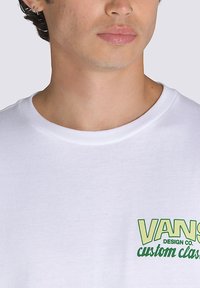 Camiseta blanca de algodón con un gráfico verde que dice "VANS DESIGN CO. custom classics" en la zona izquierda del pecho.