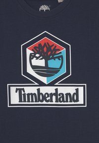 Mörkblå t-shirt i bomull med en framträdande Timberland-logotyp med ett träd inuti en hexagon. Logotypen har röda, blå och vita detaljer.
