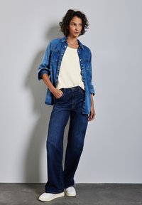 Denim skjorte med knapper over en cremefarvet top, parret med mørkeblå bell-bottom jeans og hvide sneakers. Glatte teksturer, afslappet pasform.