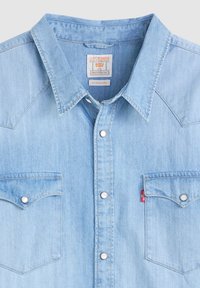 Jasnoniebieska koszula denimowa z zatrzaskami, dwiema kieszeniami na piersi, spiczastym kołnierzykiem i czerwoną metką Levi's na prawej kieszeni.