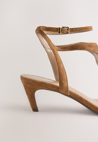 Sandale à talon en daim marron avec une fine bride à la cheville et une forme triangulaire à l'avant. Dispose d'une semelle intérieure en cuir et d'un talon court et solide.