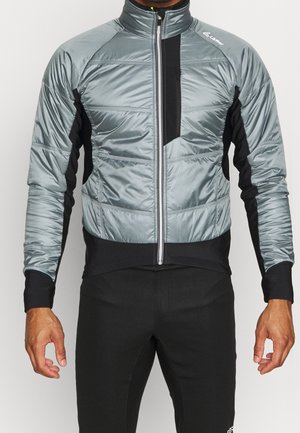 Uomo che indossa una giacca imbottita grigia e nera con cerniera frontale, abbinata a pantaloni neri, su uno sfondo chiaro e uniforme.