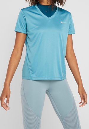 Frau trägt hellblaues kurzärmeliges Sportshirt mit V-Ausschnitt und passende Leggings, steht vor einfarbigem Hintergrund.