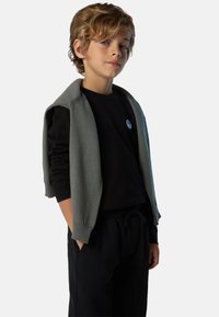 Suéter sin mangas gris sobre una camisa de manga larga negra, combinado con pantalones negros. Presenta un pequeño logotipo azul en la camisa.
