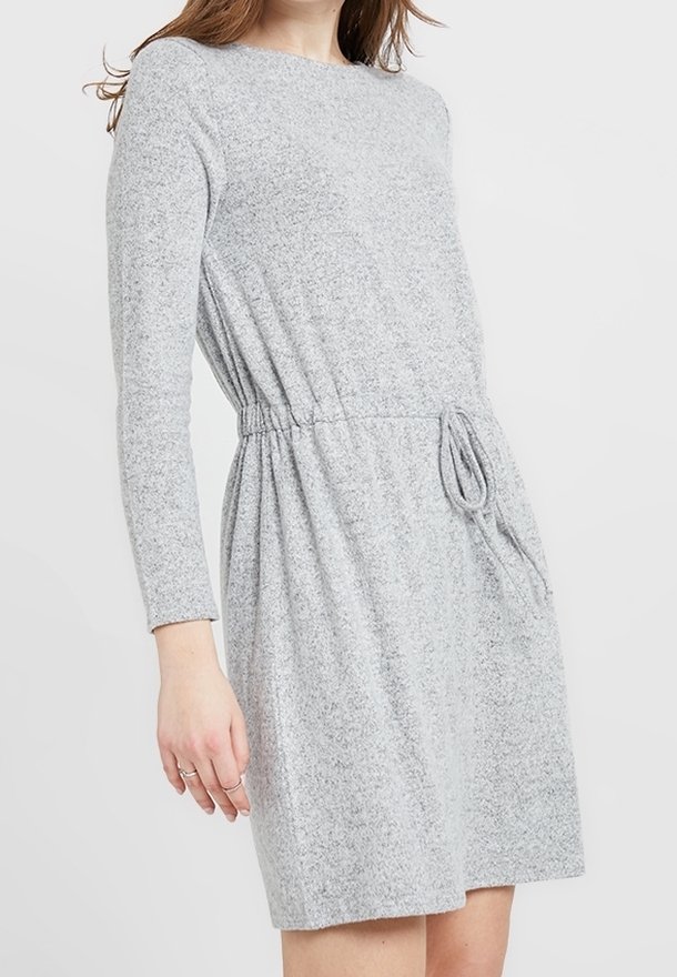 Femme portant une robe en maille gris clair à manches longues avec une taille cintrée et un détail noué sur un fond uni.