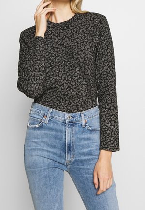 Femme portant un haut noir à manches longues avec un imprimé léopard argenté, rentré dans un jean taille haute bleu clair, main près du visage.