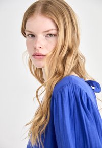 Plava bluzica s dugim, nabranim rukavima i detaljem vezivanja straga. Model ima valovitu, kosu dugine do ramena i prirodan make-up.
