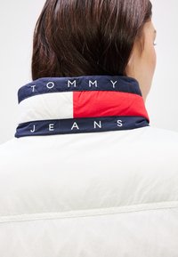 Balta striukė su tamsiai mėlynu apykakle, puošta raudonai-baltomis juostelėmis. Apykaklėje baltu siuvinėjimu yra užrašyta "TOMMY JEANS".