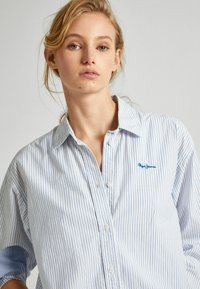 Pruhovaná košeľa s button-up zapínaním v svetlo modrej a bielej farbe, obsahujúca golier, dlhé rukávy a malé modré logo na hrudi. Jemná textúra látky.