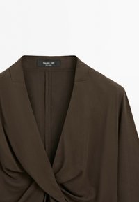 Hnědá halenka Massimo Dutti s hlubokým výstřihem do V a překříženým detailem vpředu, detailně zobrazena na čistě bílém pozadí.