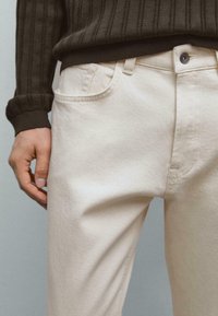 Person in cremefarbigen Jeans und einem dunklen gestreiften Pullover, mit einer entspannten Hand an der Seite vor einem schlichten Hintergrund.