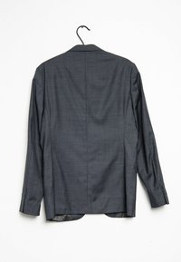 Tmavě šedý oblekový kabát vyrobený z texturované látky, se speciálním rozparkem, přiléhavým střihem a knoflíkovými detaily na rukávech. Visí na černém háčku.