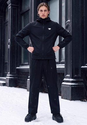 Jeune personne debout avec assurance sur un trottoir enneigé, portant une veste à capuche noire, un pantalon et des bottes devant une façade de bâtiment sombre et détaillée.