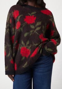 Maglione lavorato a maglia in tessuto scuro con ampio motivo floreale rosso e dettagli verdi. Presenta un ampio scollo e polsini a coste. Abbinato a jeans scuri.