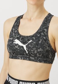 Μαύρο σπορ σουτιέν με καμουφλάζ σχέδιο, με λευκό λογότυπο Puma, σχεδίαση racerback και μαλακή ελαστική ζώνη για στήριξη.