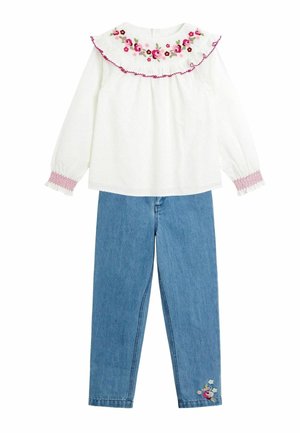 Weißes Langarmshirt mit floralem Stickerei am Kragen, kombiniert mit blauen Jeanshosen, die blumige Stickereidetails am Bein aufweisen.