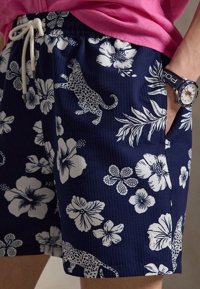 Pantalones cortos azul marino con estampados blancos de flores y leopardo, cintura con cordón, llevados con una camisa rosa y un reloj en la muñeca derecha.